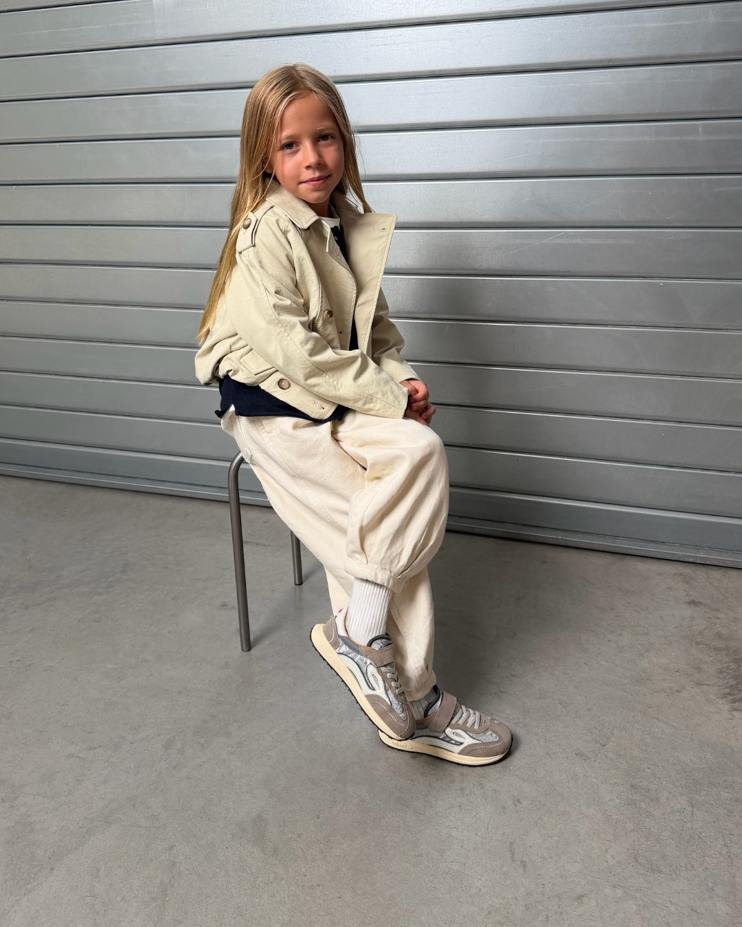 Barn med sølvgrå børnesneakers på fod, siddende på skammel foran metalport