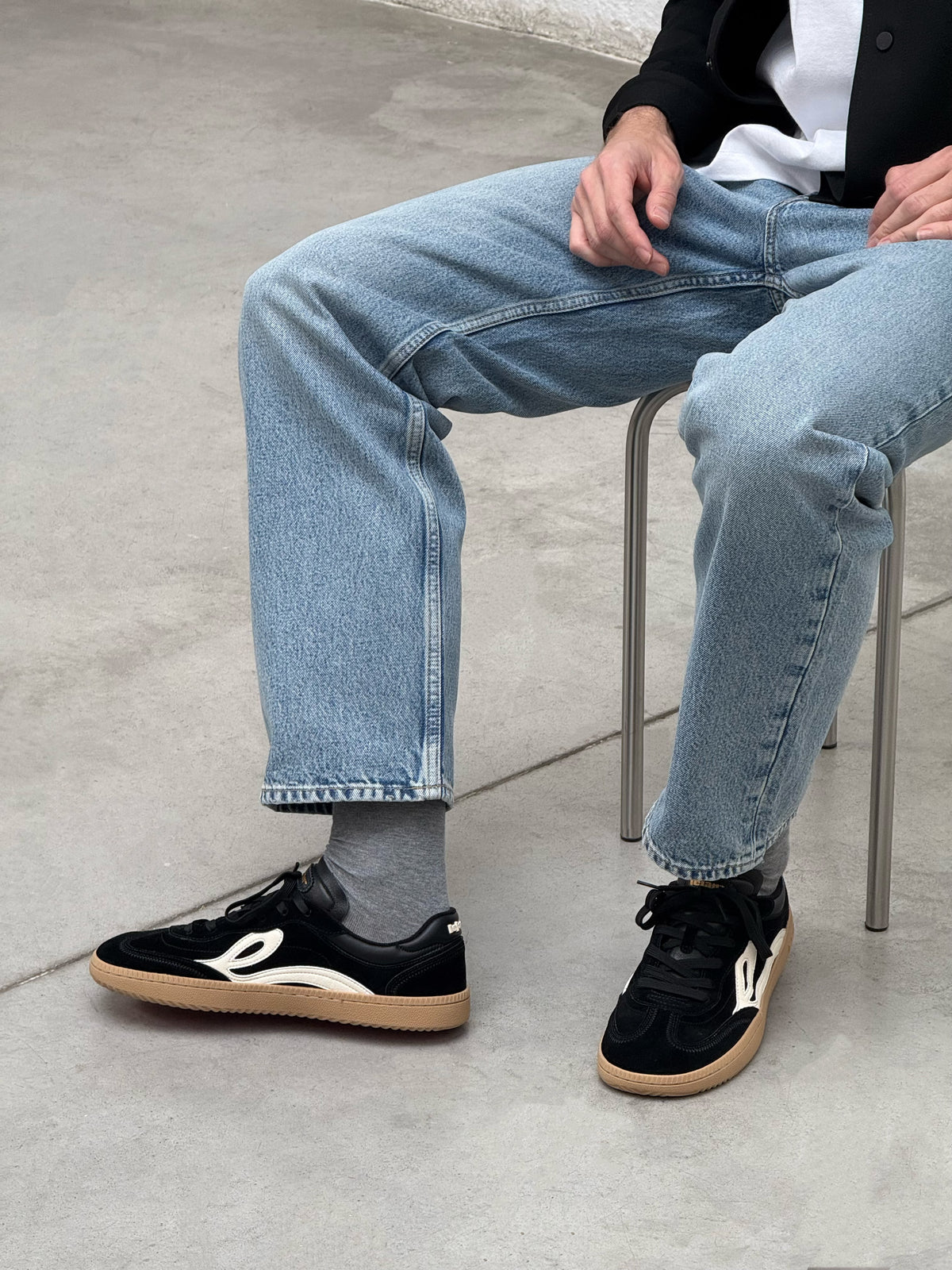 Nærbillede af sorte Lejan One på fødderne af en siddende mand i lyse jeans