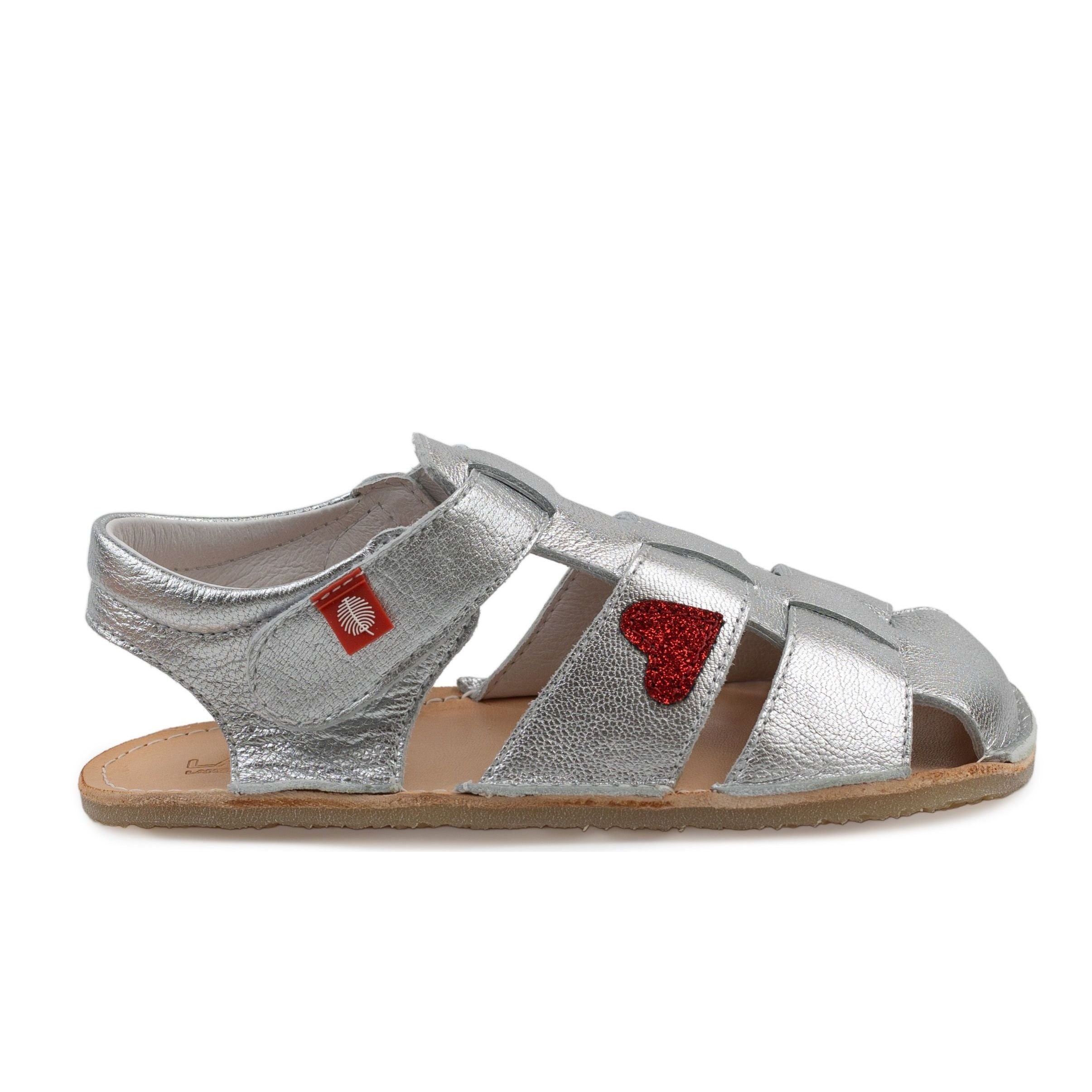 Tikki Lasta Leather Cuore bæresandal til børn i sølv med rød glitterhjerte. Unisexdesign med åben tå og velcrolukning, behagelig naturfarvet sål og mærketag.