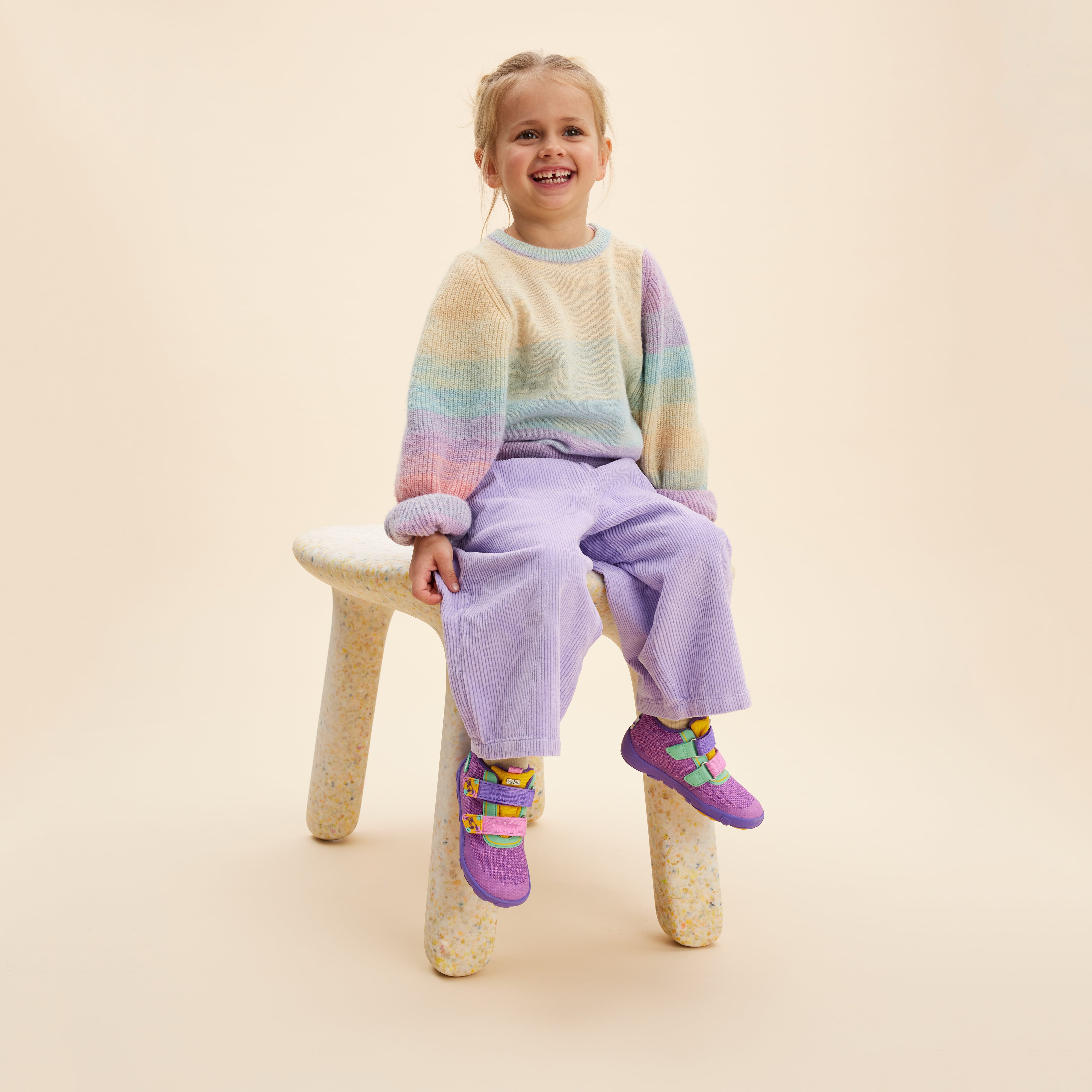 Barn iført lilla Affenzahn Lowboot Knit Happy Creative Toucan sko, med grønne og gule detaljer, sidder på en stol, i farvestrålende tøj. Barfodssko til børn i en kreativ stil.