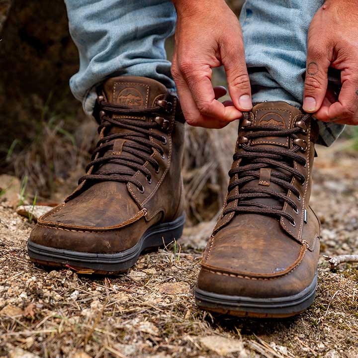 Lems Boulder Boot Grip Waterproof Dakota på en model i en udendørs naturindstilling, brune læderstøvler med mørkebrune snørebånd og højt skaft velegnet til udendørs aktiviteter.