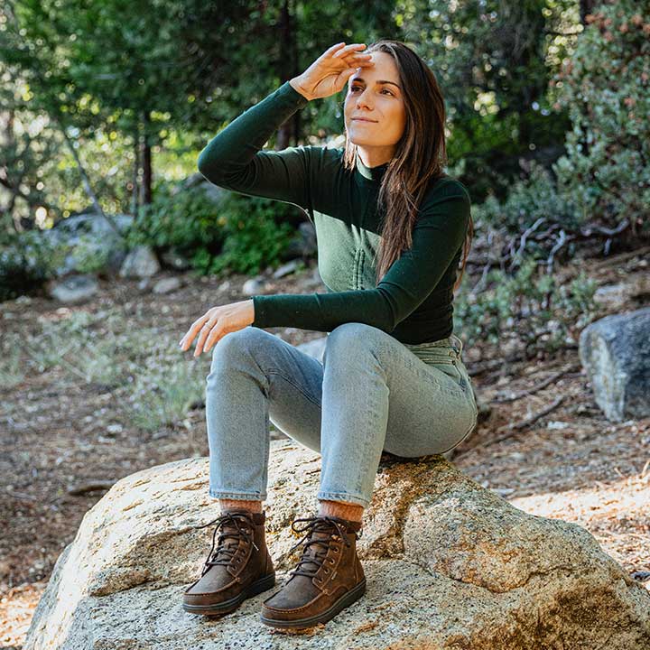 Person iført Lems Boulder Boot Grip Waterproof i Dakota variant, brun læder, siddende på sten i udendørs naturmiljø. Støvlerne er karakteriseret ved robuste syninger og mørke snørebånd.