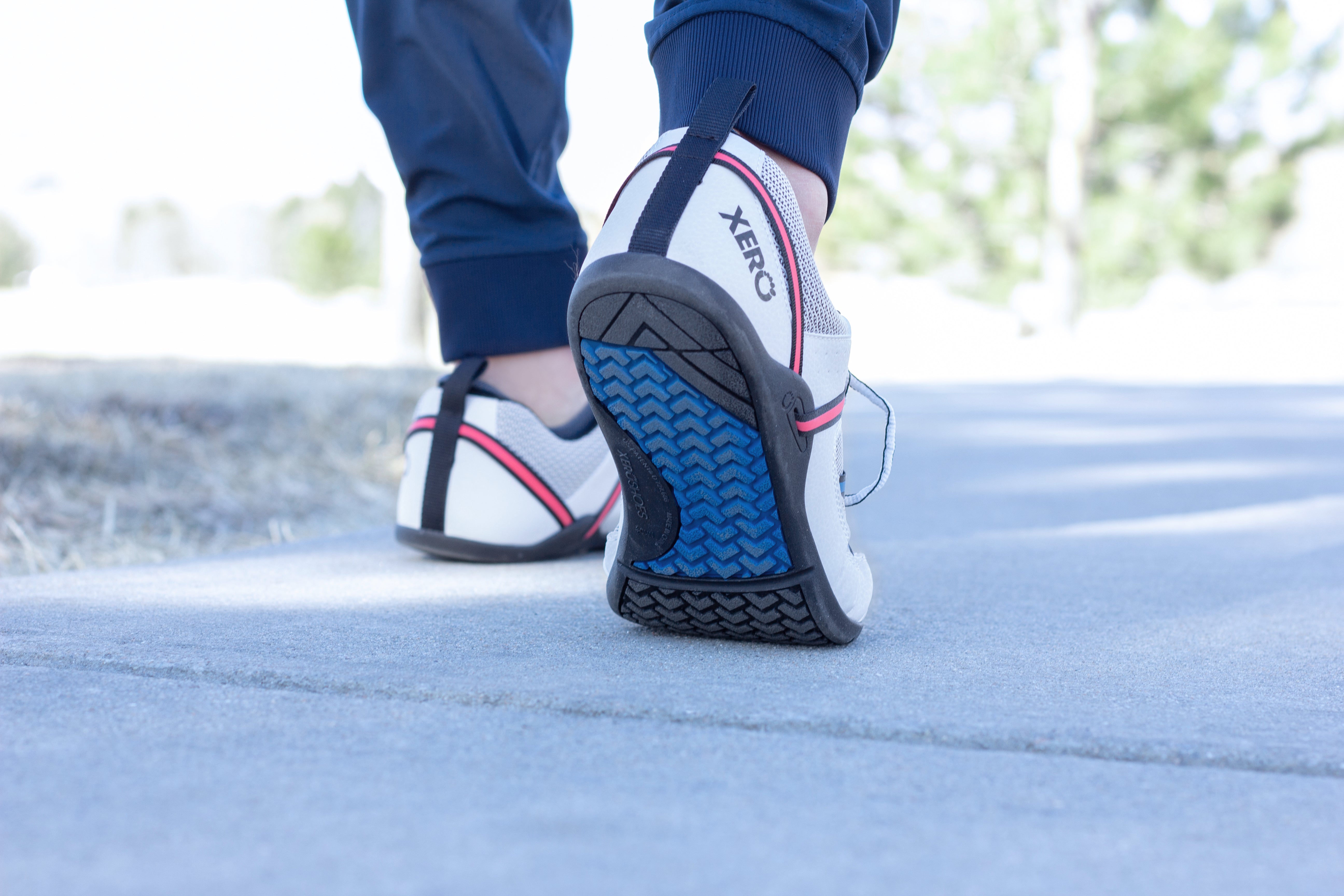 En mand går iført Xero Shoes Prio Men i Lunar-varianten, en minimalistisk barfodssko med reflekterende stropper og blå sål for naturlig bevægelse. Lavet af veganske materialer.