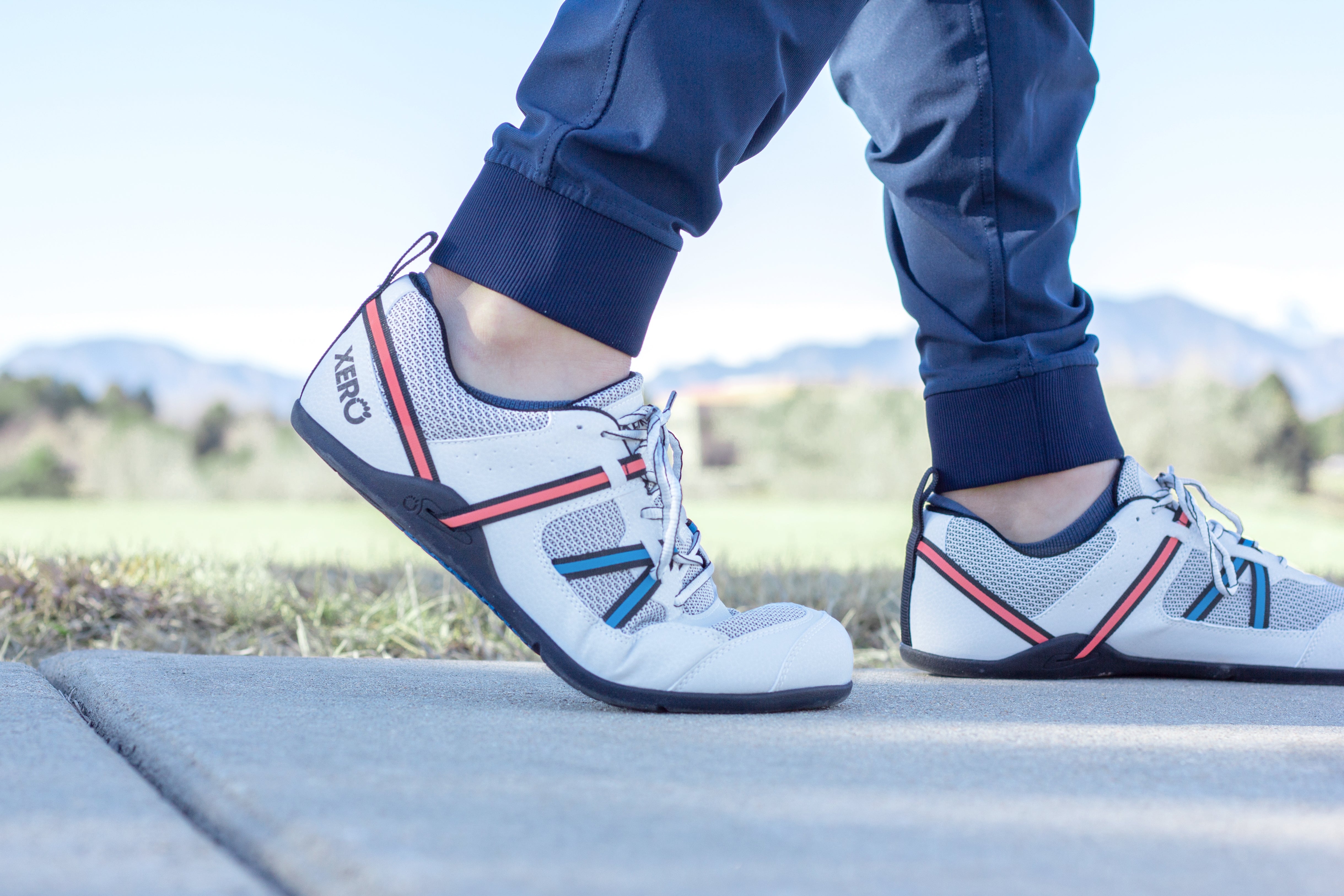 Xero Shoes Prio Men Lunar variant worn outdoors. White mesh and synthetic upper with pink and blue straps, thin black sole, highlighting flexible barefoot design.